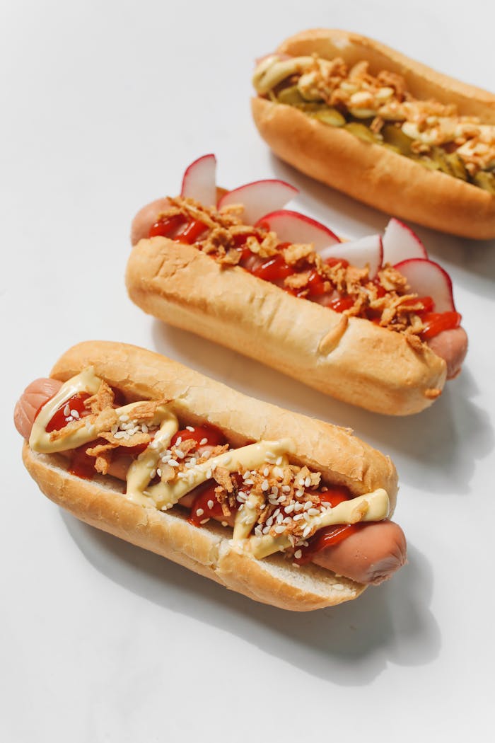 Three gourmet hot dogs topped with mustard, ketchup, and fresh vegetables on a white background.